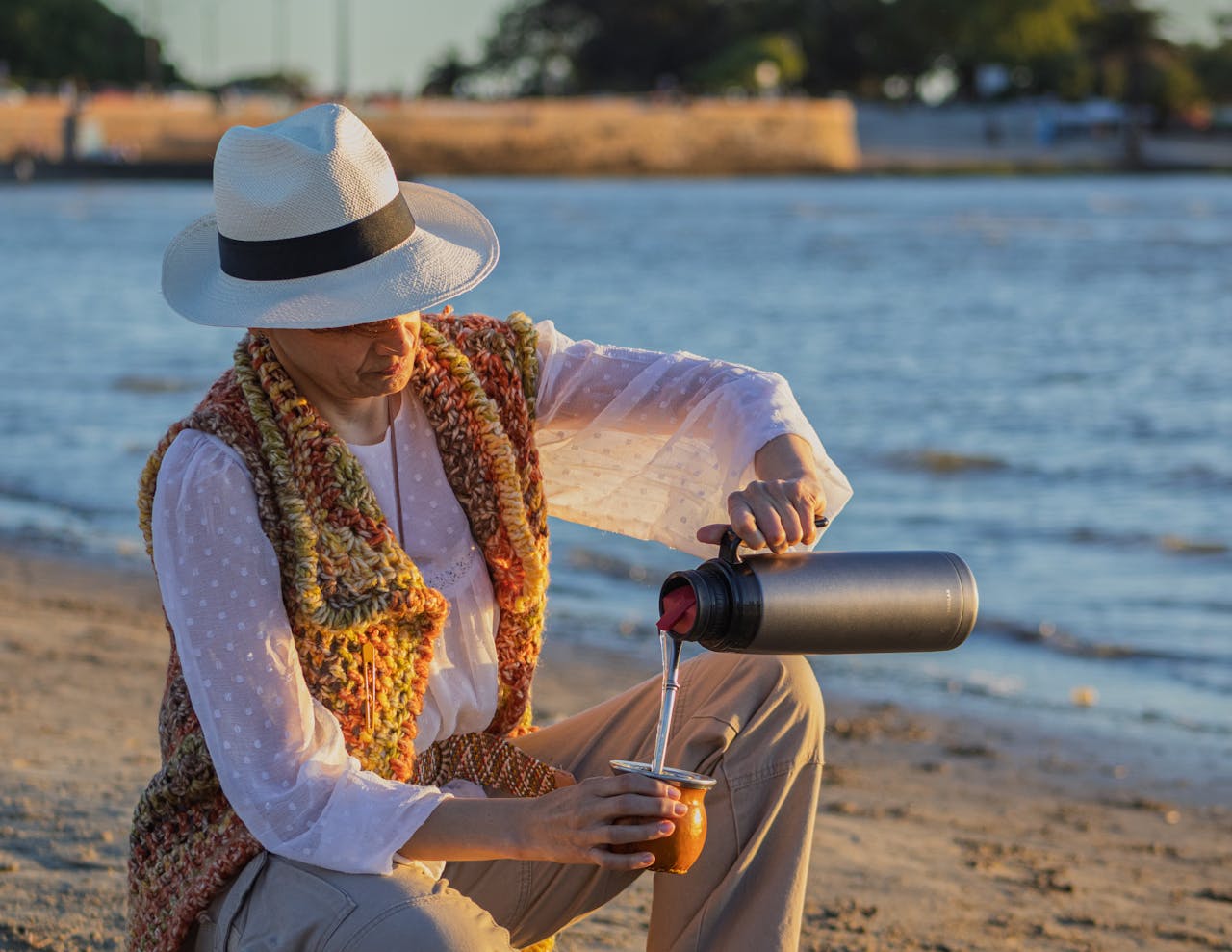 Mastering the First Impression: Your intriguing post title goes here Adult woman pours yerba mate by the beach in Montevideo, Uruguay, wearing a hat and colorful vest.